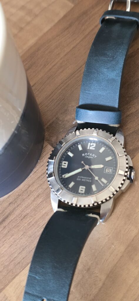 Rotary dive watch with a black dial, Arabic numerals at 12, 6, and 9, luminous markers, a date window at 3 o'clock, a polished steel bezel, and a blue leather strap resting on a wooden surface.