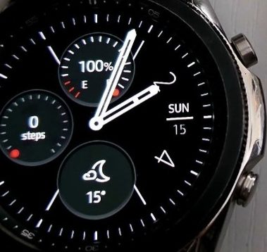 Close-up of a Samsung Galaxy smartwatch face displaying a digital dial with analog hands, featuring sub-dials for battery percentage, step count, and weather alongside a date window reading Sunday the 15th.