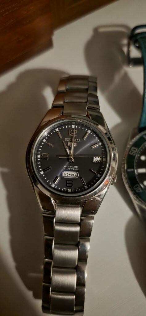 Seiko 5 automatic watch with a dark grey sunburst dial, featuring silver Arabic numerals at 6, 9, and 12, a day-date window at the 4 o'clock position, and a steel bracelet.