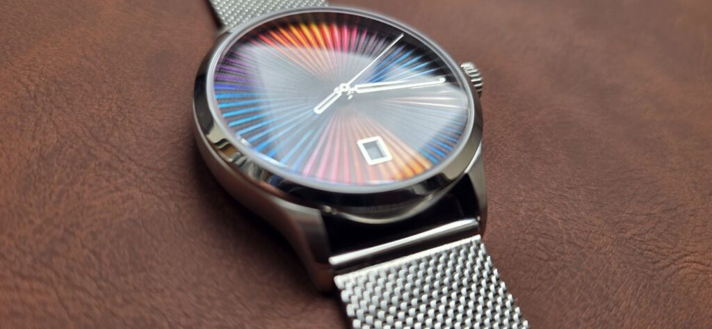 Review: Mezei First Anniversary Edition Low-angle close-up shot of the Mezei First Anniversary Edition watch showing the domed crystal reflection over its rainbow sunburst dial, polished stainless steel case edge, and mesh bracelet on a brown leather surface.