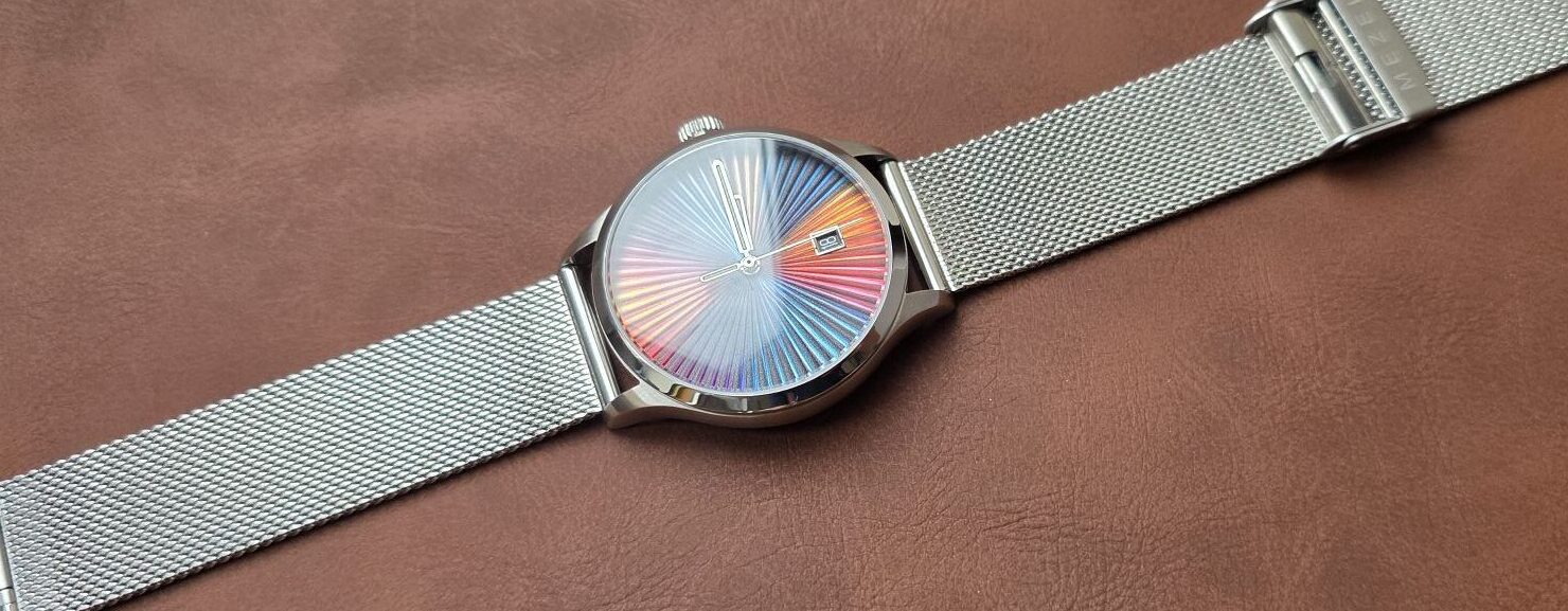 Review: Mezei First Anniversary Edition Angled view of the Mezei First Anniversary Edition watch with a stainless steel mesh strap laid flat across a smooth brown leather background, displaying its colorful rainbow sunburst dial and date display.