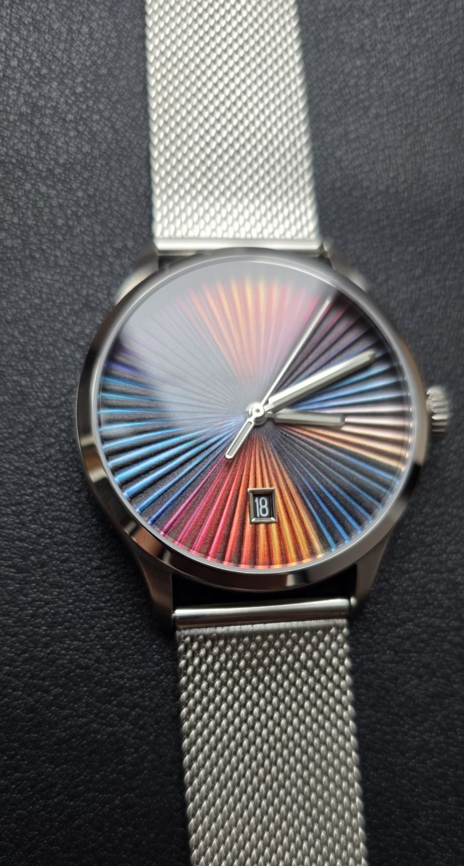 Review: Mezei First Anniversary Edition Vertical close-up shot of the Mezei First Anniversary Edition watch highlighting the intricate rainbow sunburst dial texture, skeletonized silver hands, 6 o'clock date window, and stainless steel mesh bracelet against a black leather background.