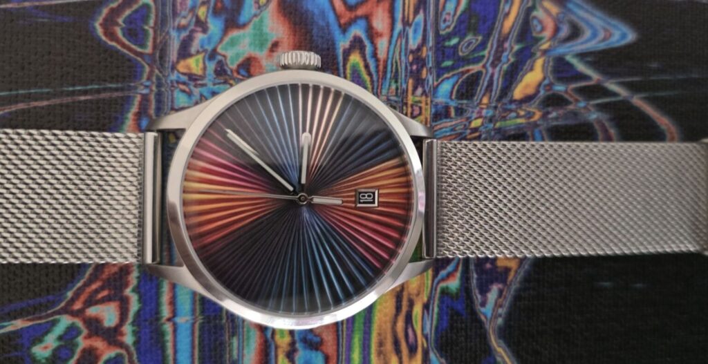 Review: Mezei First Anniversary Edition Horizontal flat lay photo of the Mezei First Anniversary Edition watch with a stainless steel mesh strap, laid across a trippy, psychedelic multicolored patterned surface that complements the rainbow sunburst dial.