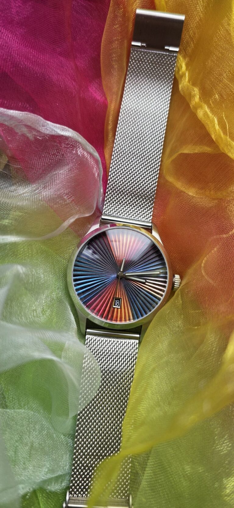 Top-down portrait shot of the Mezei First Anniversary Edition watch with mesh bracelet, resting on vibrant multicolored flowing silk fabric that mirrors the rainbow sunburst dial design.