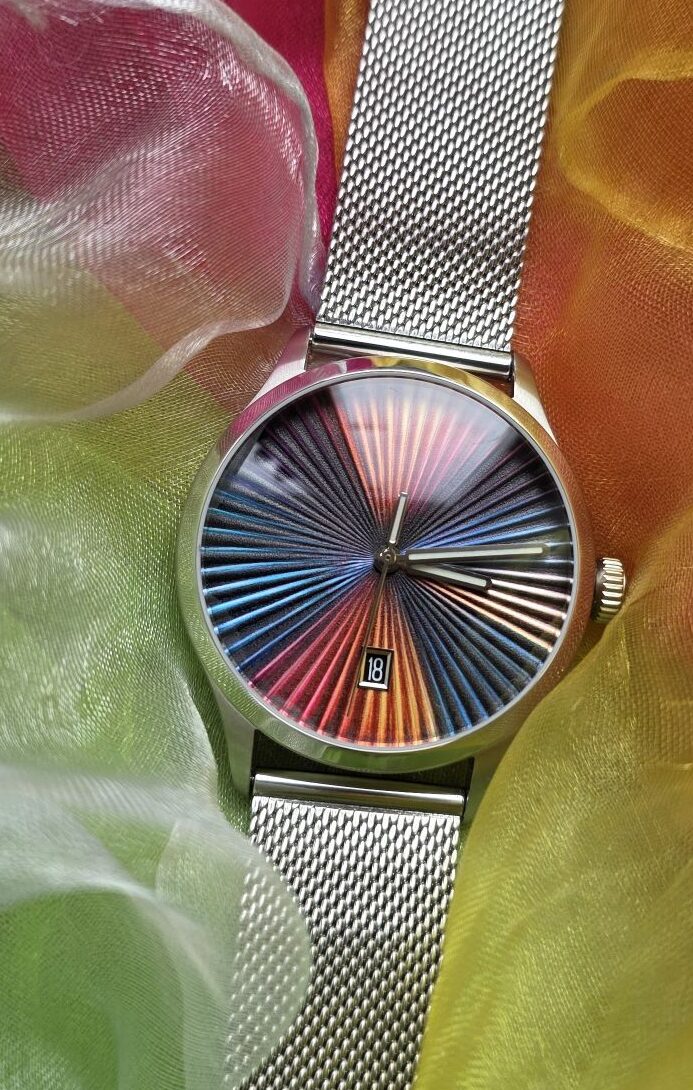 Top-down portrait shot of the Mezei First Anniversary Edition watch with mesh bracelet, resting on vibrant multicolored flowing silk fabric that mirrors the rainbow sunburst dial design.