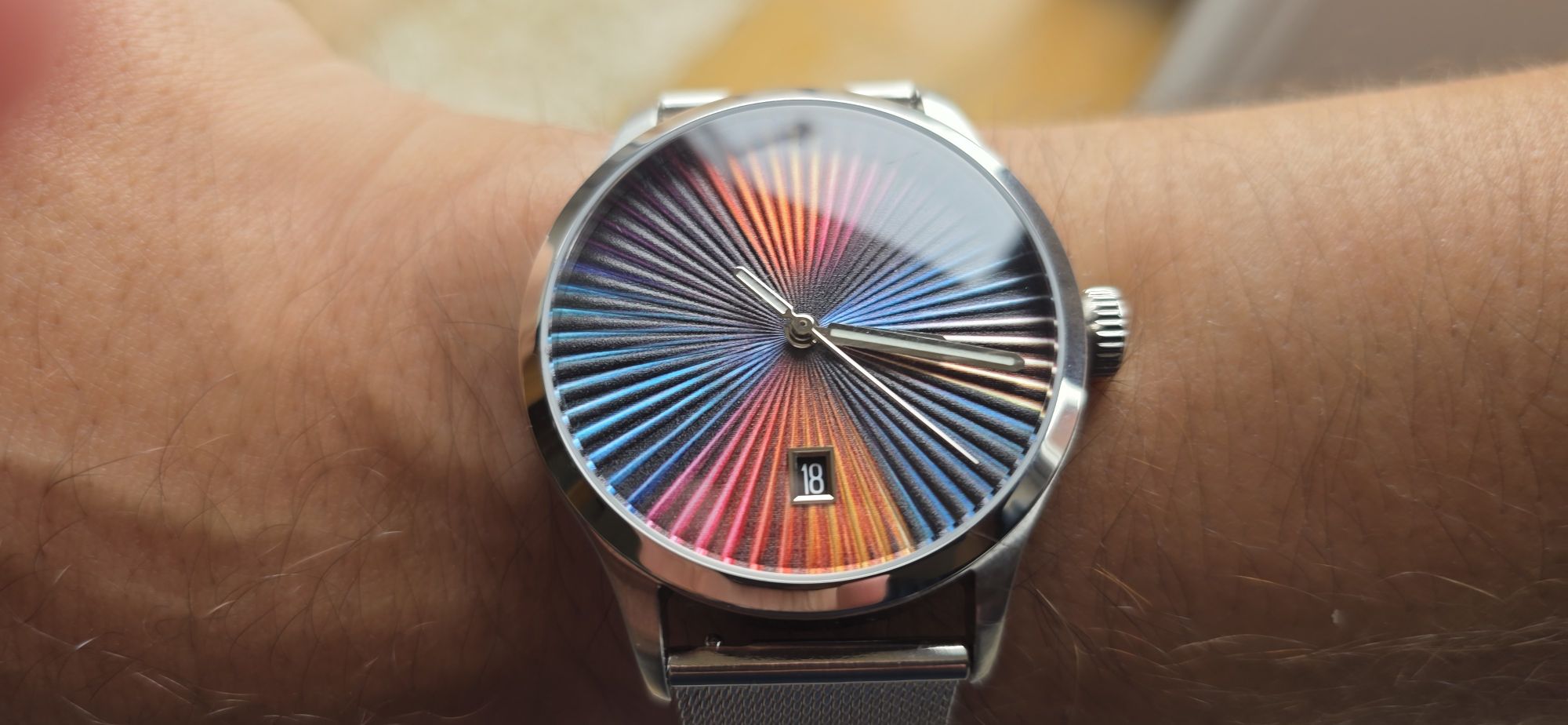 Review: Mezei First Anniversary Edition Direct wrist shot of the Mezei First Anniversary Edition watch on a stainless steel mesh bracelet, displaying its vibrant rainbow sunburst textured dial and date window at 6 o'clock under natural lighting.