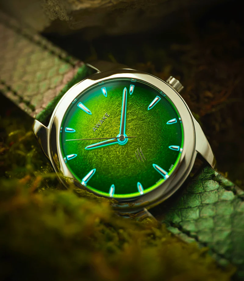 Awake Watches Close-up of an Awake Concept Jurassic edition watch with a glowing green textured dial, luminescent markers, and a matching green scales leather strap resting on a natural mossy background.