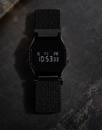Overhead flat lay studio shot of an Eight digital watch featuring an octagonal black case, negative LCD digital display, and textured black strap resting on a dark slate background.