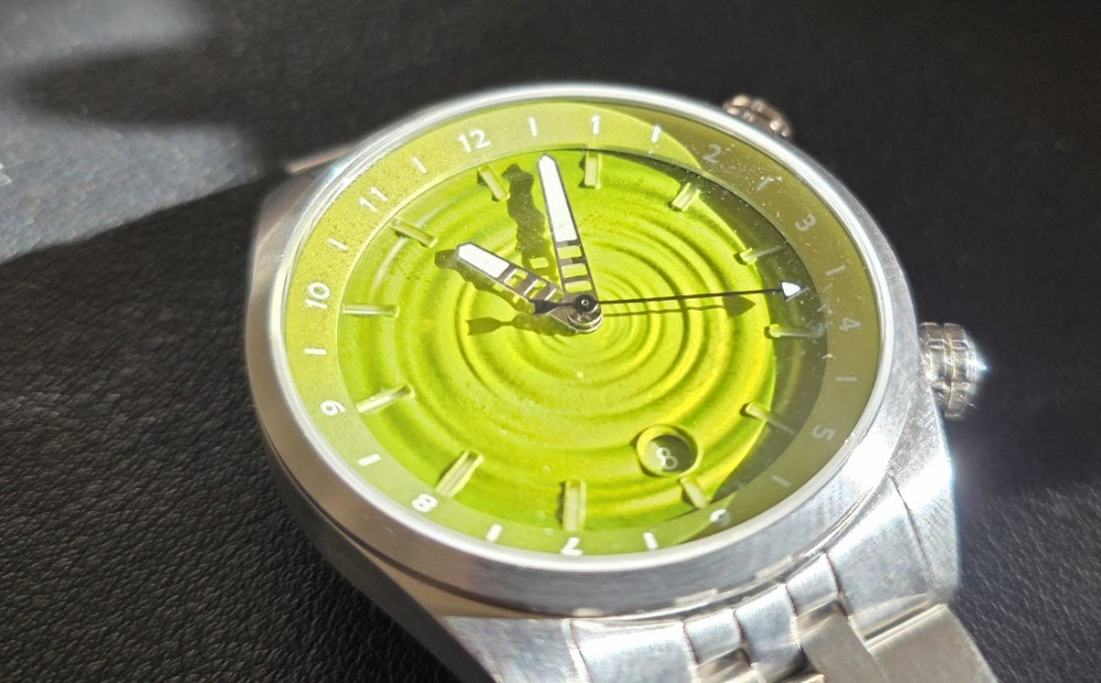 Close-up angled view of The Skipping Stone by Golby watch resting on a dark surface, displaying the light reflections across the green water-ripple dial and dual crowns