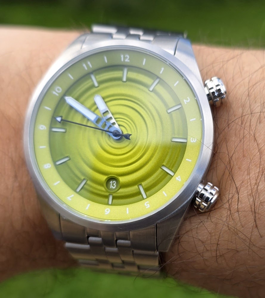 On-wrist close-up photo of The Skipping Stone by Golby automatic watch showing the lime-green concentric ripple dial, skeletonized hands, dual crowns, and stainless steel bracelet.