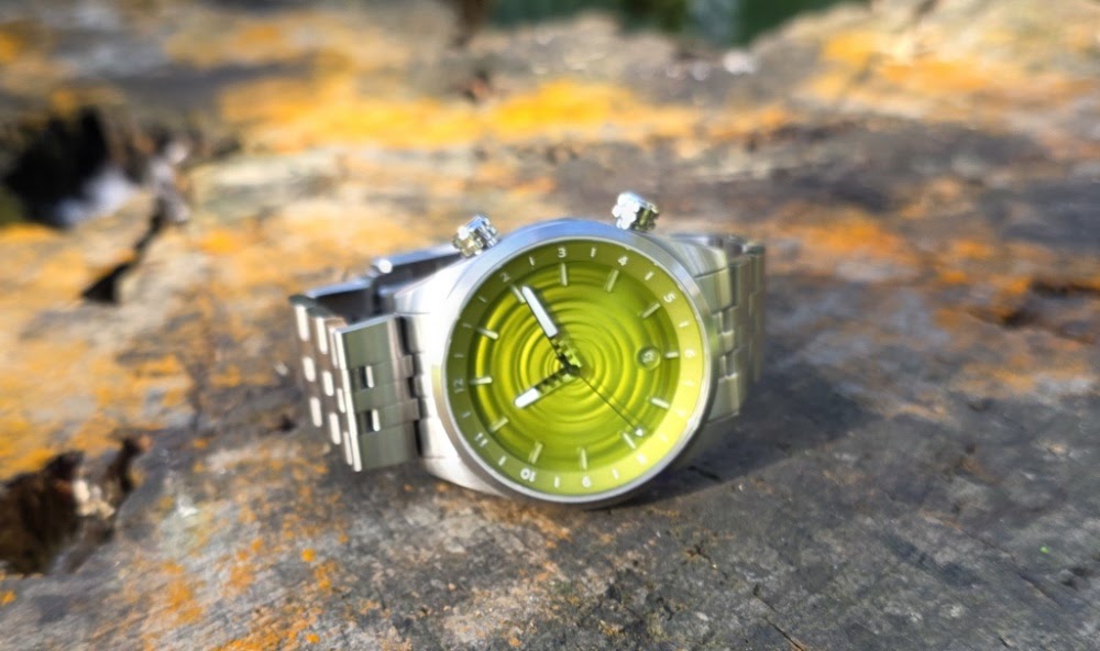 The Skipping Stone by Golby watch with stainless steel bracelet resting horizontally on a textured wood surface, highlighting its lime-green ripple dial and dual crown setup.