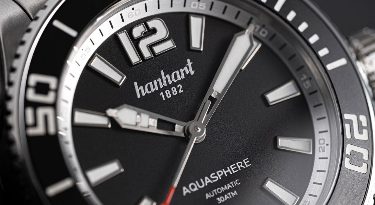 Macro close-up shot of the Hanhart Aquasphere Free Diver automatic watch featuring a matte black dial, bold applied hour indices, skeletonized hands, red-tipped seconds hand, and a knurled dive bezel.