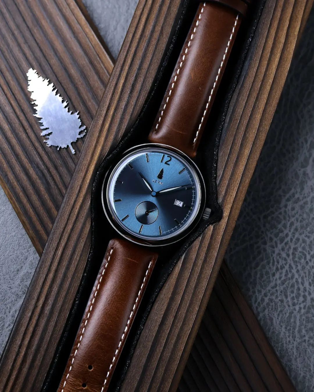 Le Pin Watches Top-down view of a Le Pin blue dial watch with a brown leather strap resting inside a slotted dark wooden presentation box alongside a silver pine tree emblem.