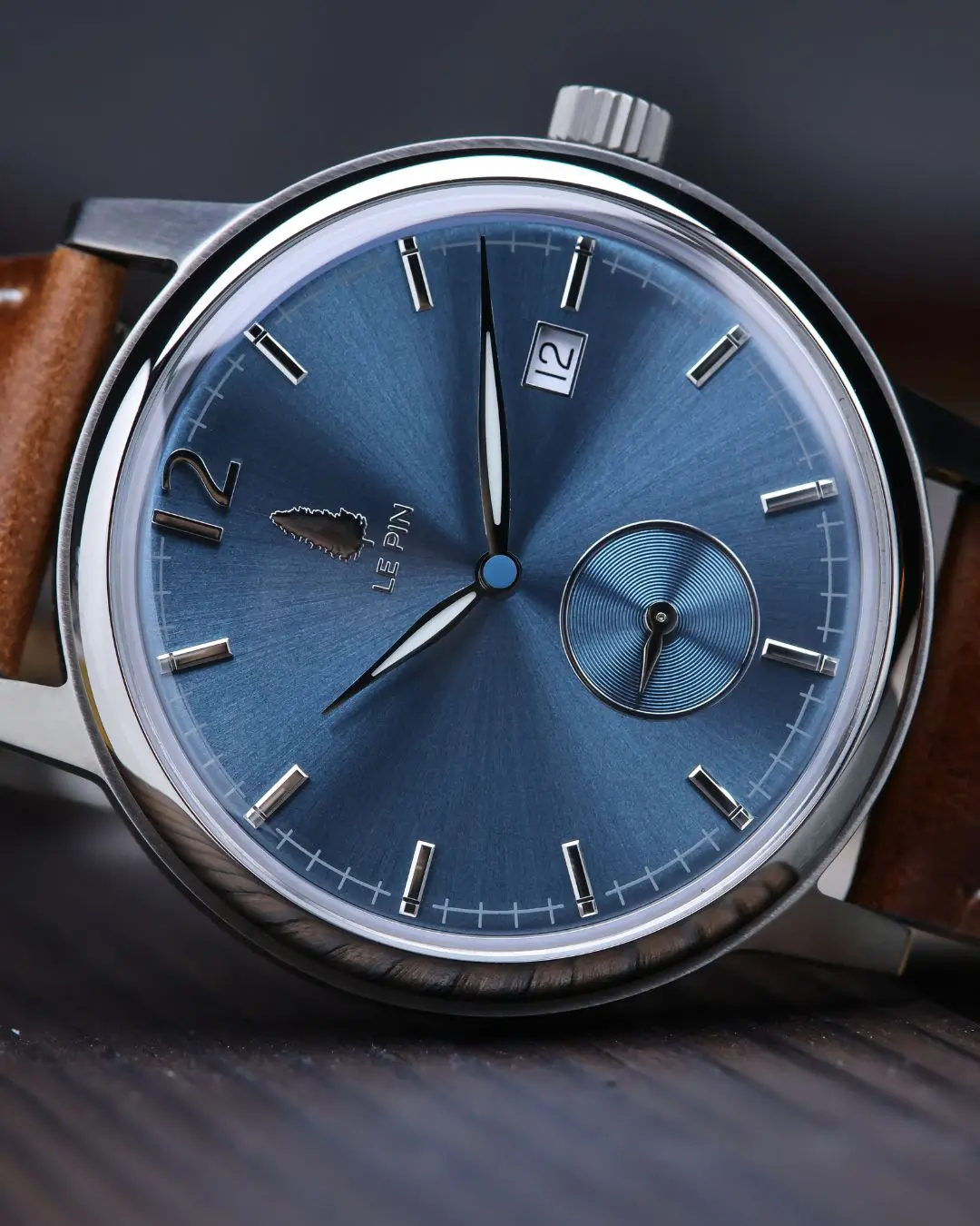 Le Pin Watches Macro close-up shot of a Le Pin dress watch featuring a blue sunburst dial, pine tree logo emblem, small seconds subdial, date window at 2 o'clock, and a brown leather strap.