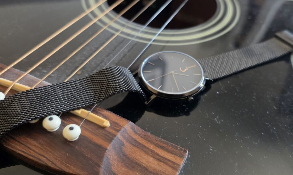 Luthier Luthier LP Black watch with a mesh bracelet lying across the bridge and strings of a black acoustic guitar.
