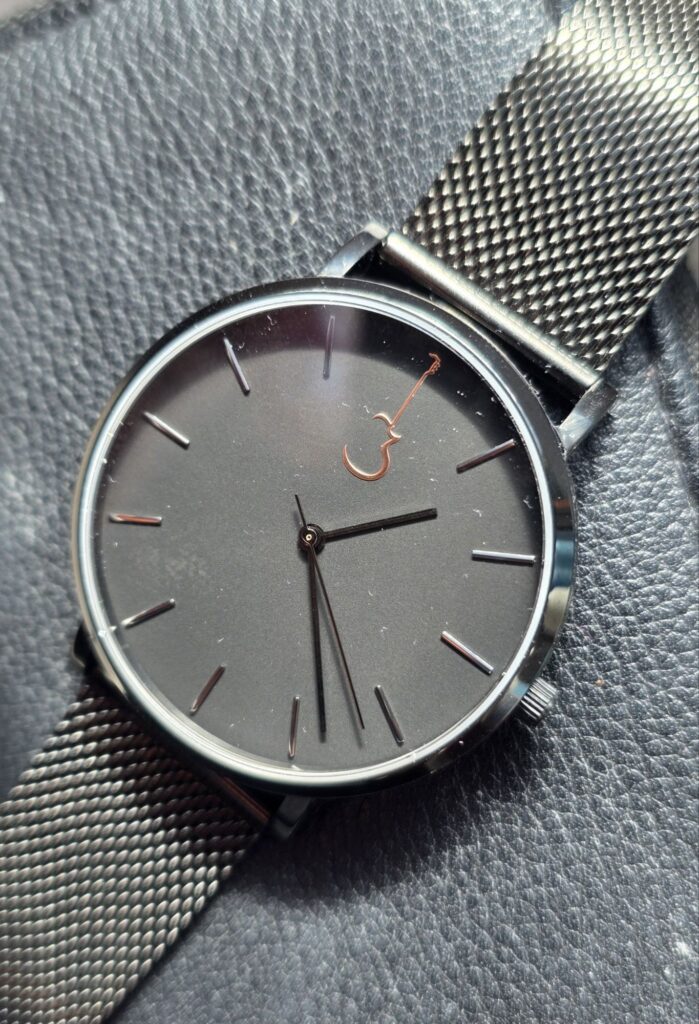 Luthier Macro close-up of the Luthier LP Black watch face highlighting the rose gold Les Paul guitar logo at 12 o'clock and the black mesh strap.