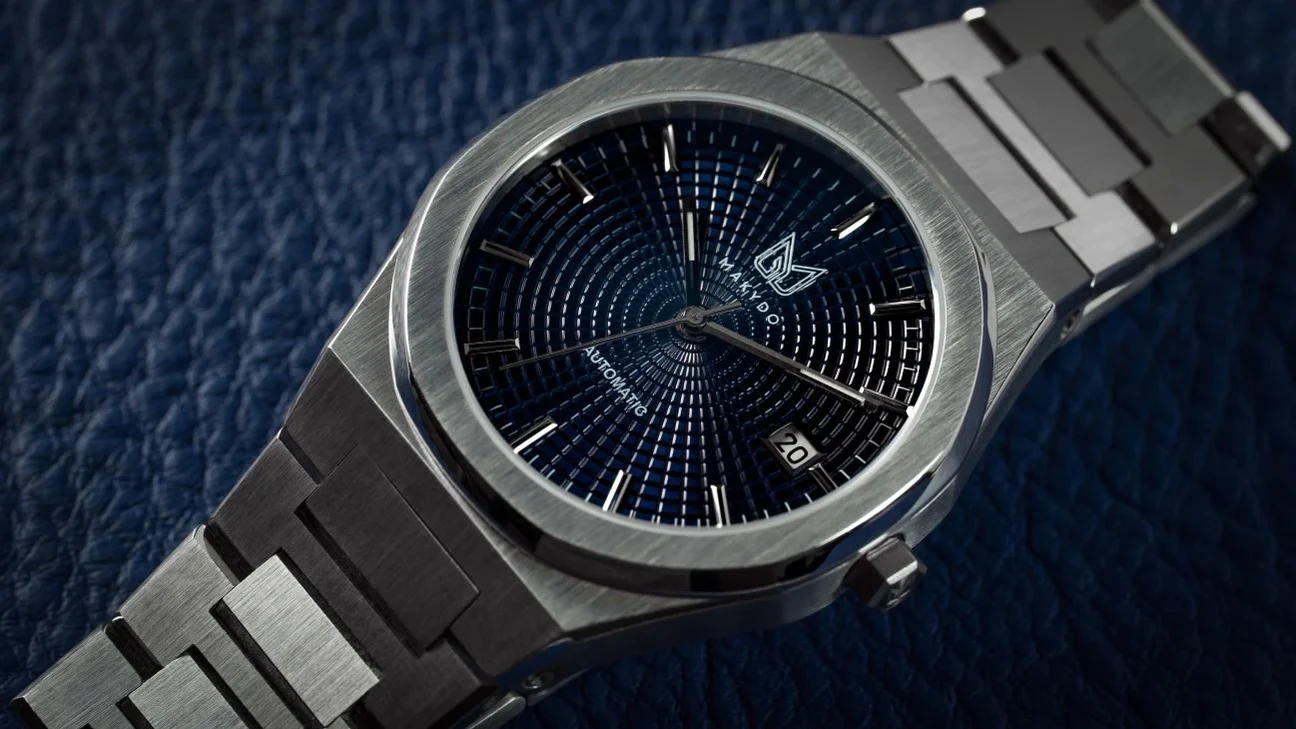 Makydo Watches Close-up macro view of the Makydo automatic watch showcasing its intricate blue textured dial, date window at 4:30, integrated brushed stainless steel bracelet, and polished bezel against a textured blue leather background.