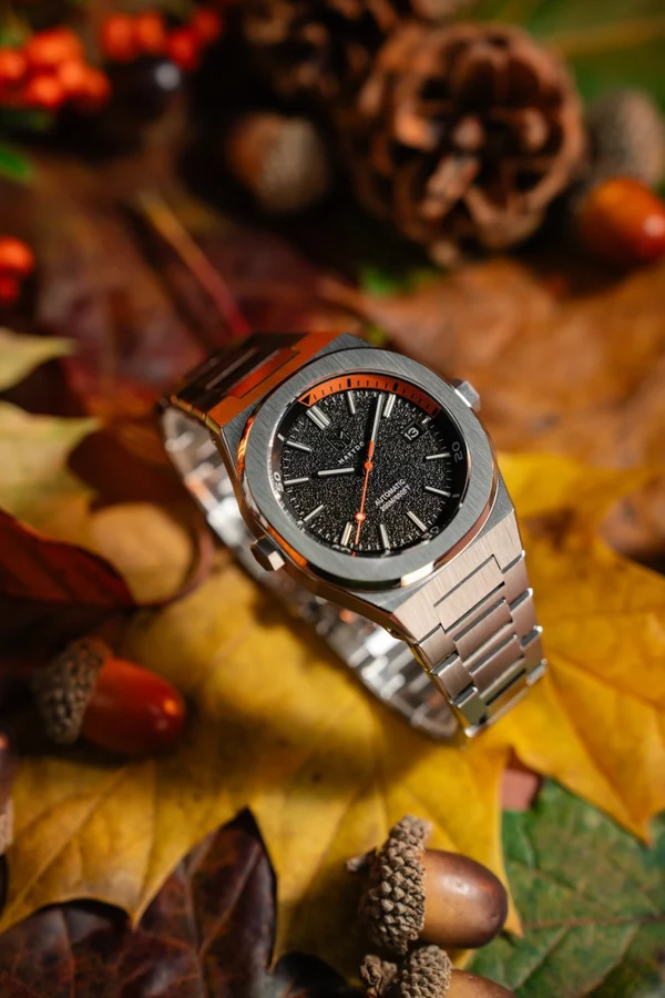 Makydo Watches High-angle macro lifestyle shot of the Makydo automatic tool watch featuring a textured black dial, orange chapter ring accents, and integrated steel bracelet, resting on a rustic bed of autumn leaves and acorns.
