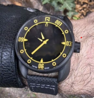 Protocol Watches Close-up wrist shot of a Protocol watch in a black PVD case with a dark textured dial, high-visibility yellow hands and chapter ring markings, and a black fabric strap.