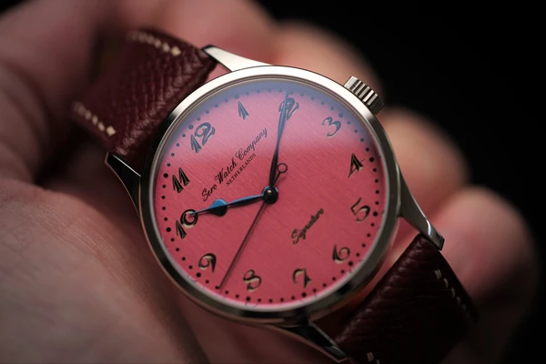 Sero Watch Company : A Sero Watch Company Signature wristwatch held in hand, featuring a vibrant brushed pink dial, blued steel hands, black Arabic numerals, and a dark burgundy leather strap.